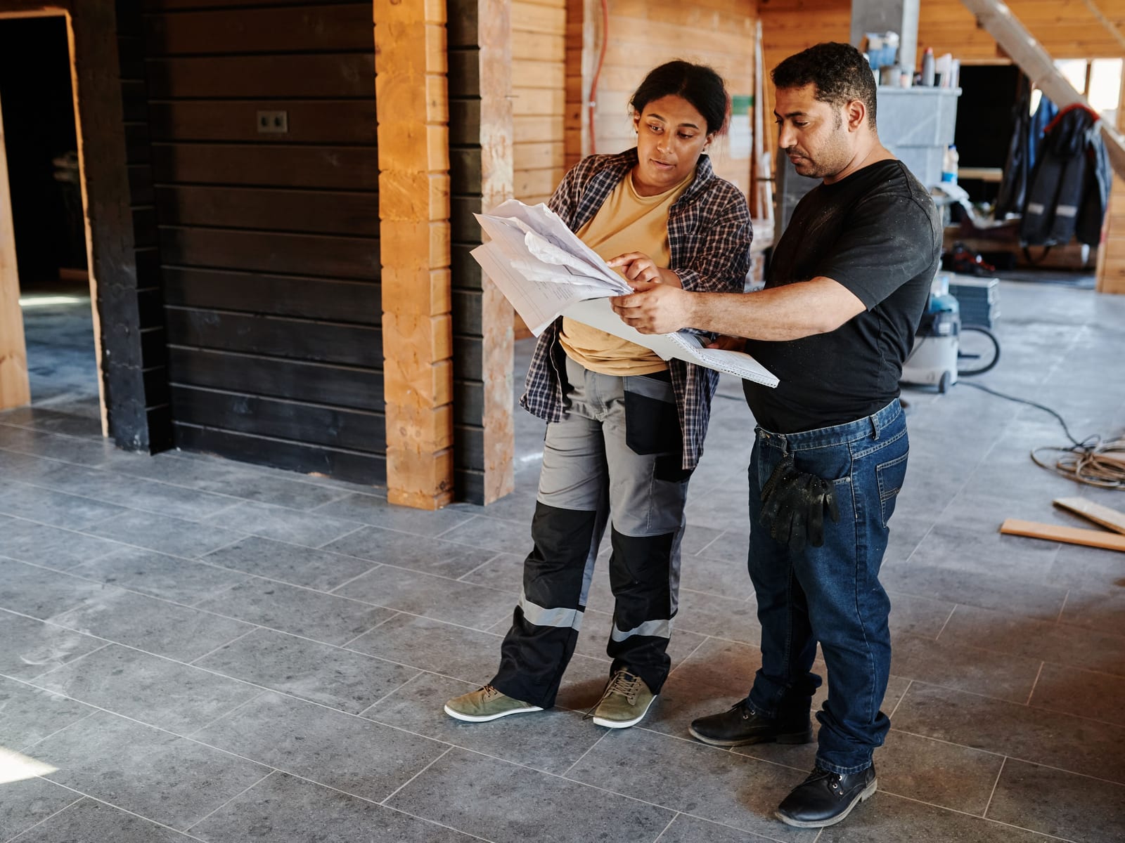Two people in work clothes standing together in an unfinished timber interior, reading through a drawing held between them.