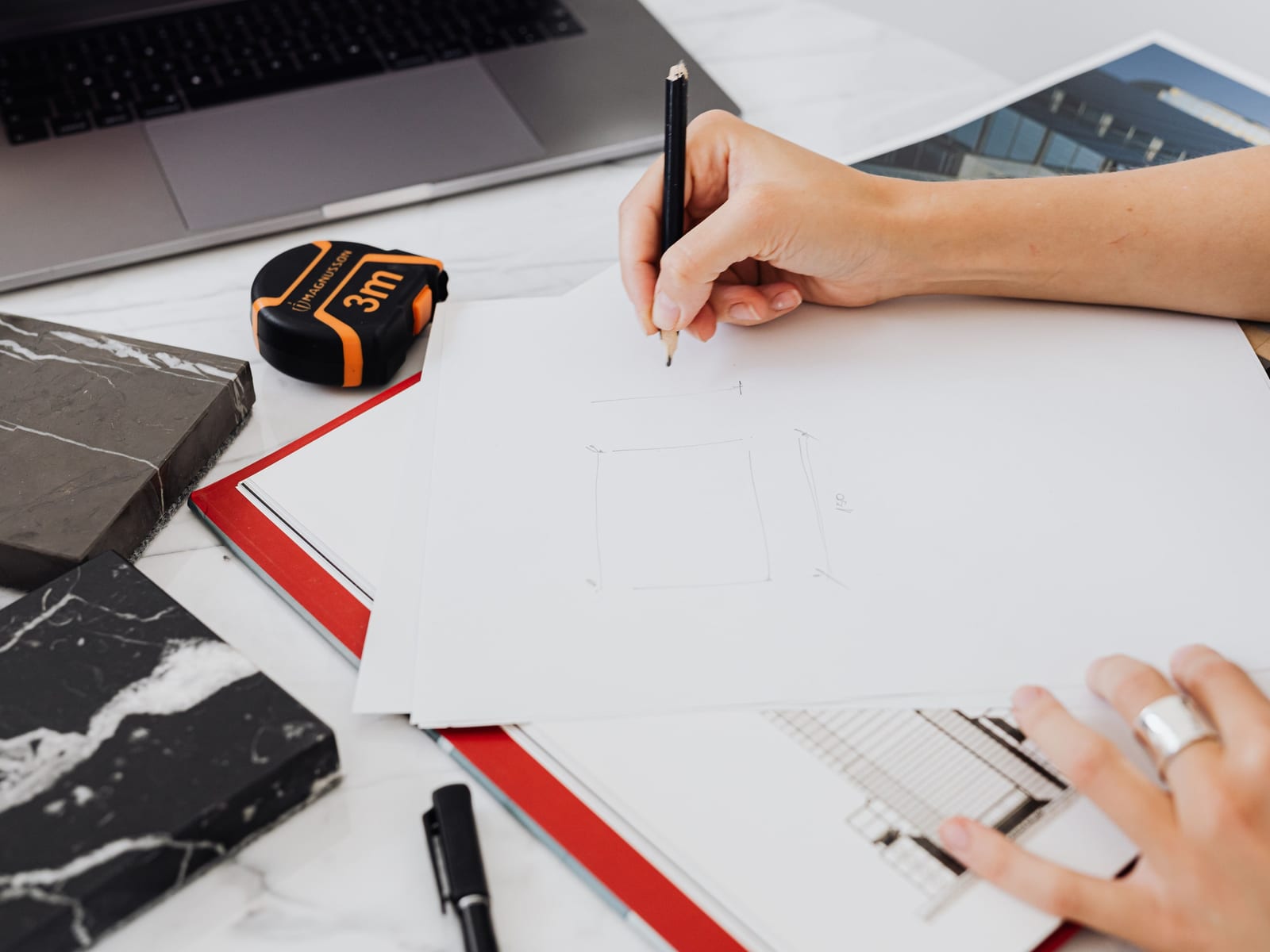 A hand sketching a room plan in pencil, a tape measure and a stone sample set out beside the drawing.