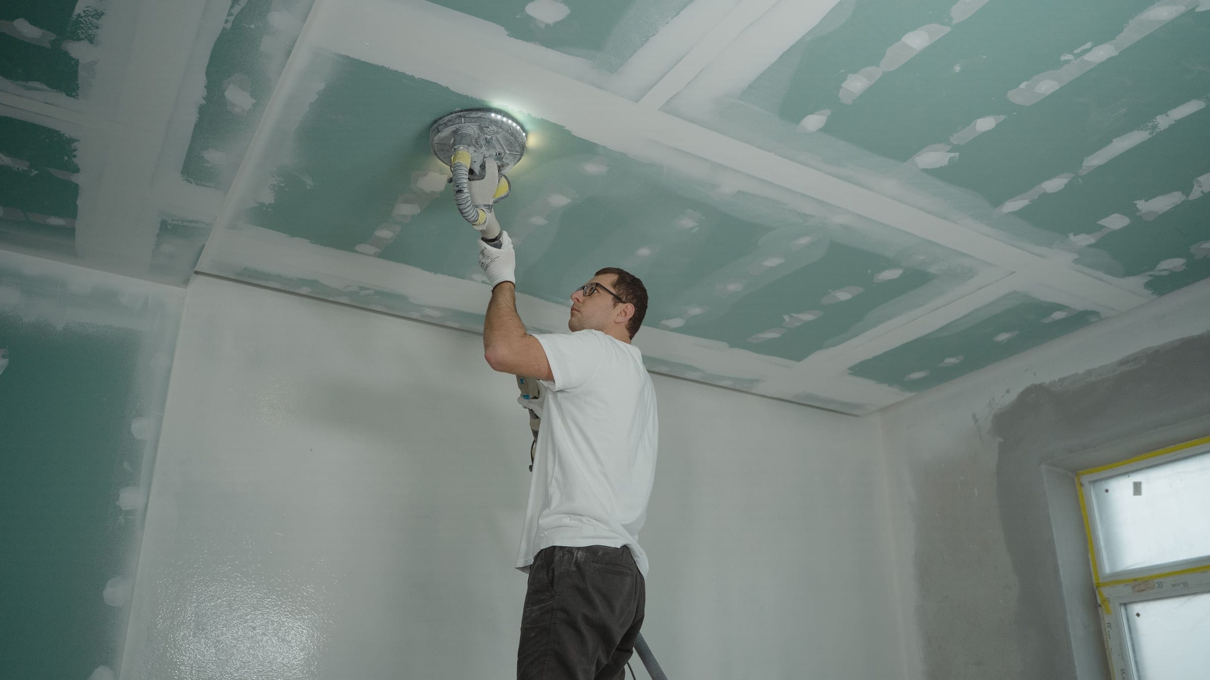 A drywall finisher on a ladder reaching overhead with a pole sander, smoothing the taped joints of a new ceiling.