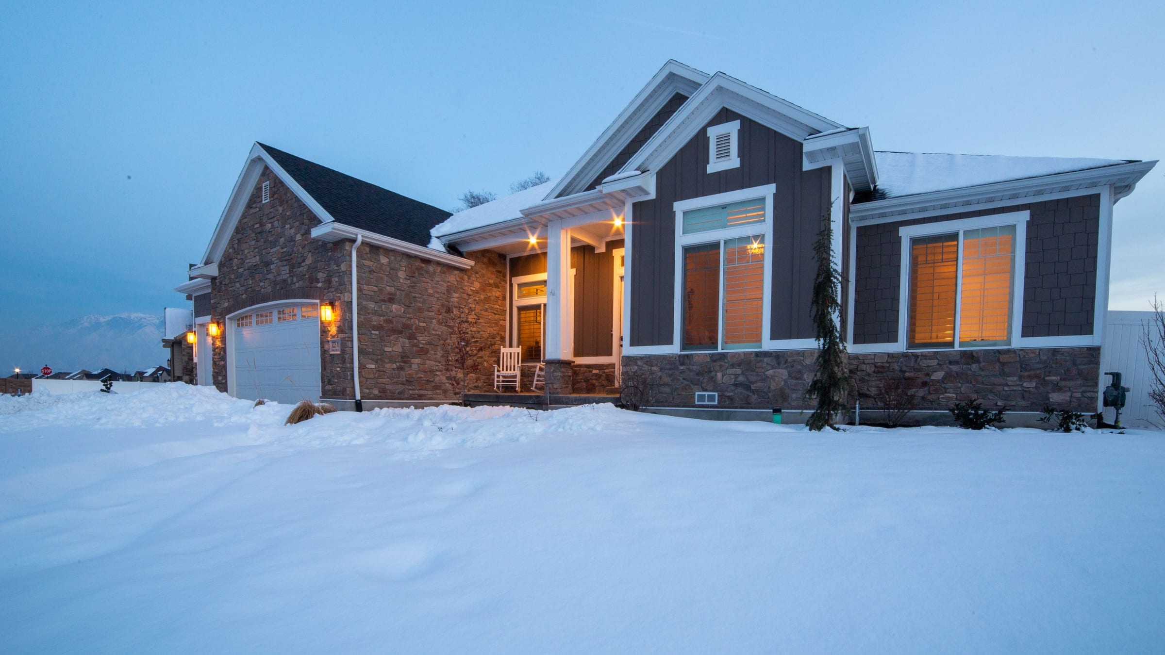 A gabled home in deep snow at blue hour, warm light in every window and a lamp lit over the porch.