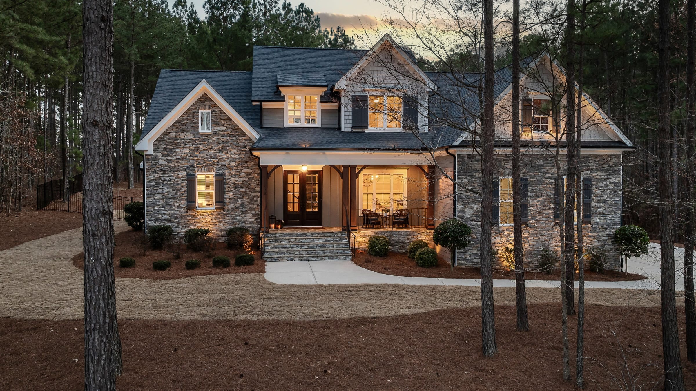 A stone and shingled home among tall pines at dusk, warm light in the windows and across the covered porch.